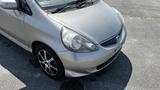 A silver 2006 Honda Fit with distinctive headlights and alloy wheels is parked with a slightly dirty exterior