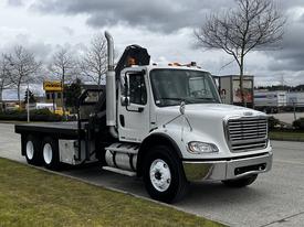 A white 2006 Freightliner M2 112 truck with a flatbed and a large exhaust stack on the driver's side, featuring orange lights on the roof and black detailing on the flatbed