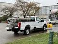 A 2023 Ford F-250 pickup truck with a flatbed and metal box attachment is parked with a yellow snow plow mounted on the front