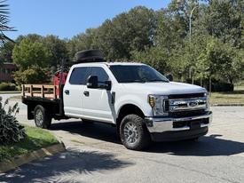 A white 2019 Ford F-350 SD with a flatbed is parked on a roadway showcasing its chrome front grille and large tires