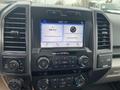 A 2019 Ford F-150 dashboard with a touchscreen display showing FM radio station and music track information along with control knobs for audio and climate settings