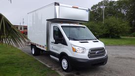 A 2018 Ford Transit box truck with a white exterior and a silver cargo box mounted on the back is parked with a visible front view