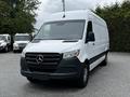 A white 2019 Mercedes-Benz Sprinter van with a black grille and front bumper is parked facing forward with its headlights on