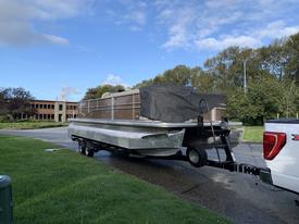 A 2013 Sylvan 13 person boat on a trailer with a cover over the seating area near a road and grassy area