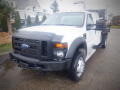 A 2008 Ford F-550 with a white exterior and a black grille parked with a flatbed truck attachment on the back