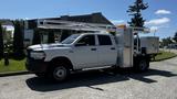 A white 2019 RAM 3500 truck with a flatbed and mounted equipment on the side, featuring a utility box and a work platform with an orange safety light