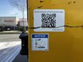 A yellow label on a truck's liftgate featuring a QR code and instructions with arrows indicating "UP" and "DOWN"