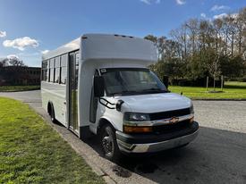 A 2015 Chevrolet Express bus with a white exterior and a large front windshield parked on a roadway with grass beside it
