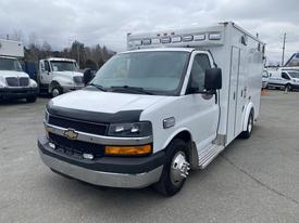 A white 2014 Chevrolet Express with a boxy shape featuring a sliding side door and emergency lights on the roof