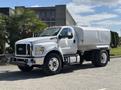 A 2016 Ford F-750 truck with a white cab and silver tank body parked on a road with a clear sky in the background
