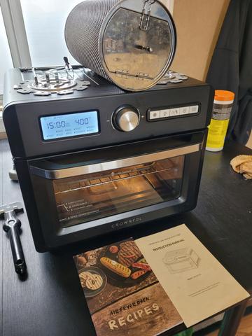 A black air fryer oven with a digital display and various buttons on the front next to a recipe booklet and a cylindrical container on top