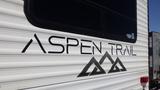 A close-up of the side of a 2022 Aspen Trail trailer featuring the name "ASPEN TRAIL" in bold black lettering with a mountain graphic below