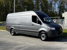 A 2021 Mercedes-Benz Sprinter van in silver with a high roof and long body design parked on a gravel surface