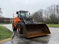 A 2018 Doosan DL300-5 wheel loader with an orange and black exterior is positioned on a wet road featuring a large bucket at the front designed for lifting and moving materials
