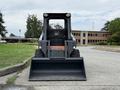 A 2017 Gehl R105 skid steer loader with a large front loader bucket positioned on a paved surface facing forward