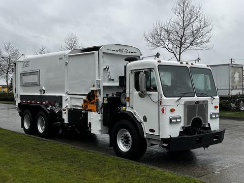 A 2010 Peterbilt 320 garbage truck with a white body and a large rear trash compartment parked on a wet surface