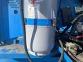 A close-up view of a gray gas cylinder with safety labels and hoses attached to blue machinery parts