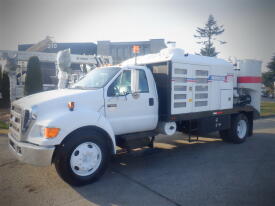 A 2007 Ford F-650 truck with a white and black body and orange beacon light on top, featuring a distinctive box-like structure in the truck bed designed for utility use