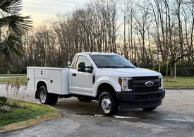 A white 2018 Ford F-350 truck with a flatbed and tool compartments on the sides is parked at an angle showing its front and side views