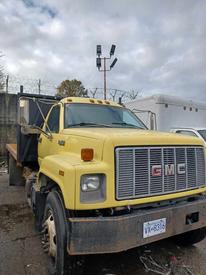 A yellow 1996 GMC C7H042 truck with a flatbed and visible rust on the wheels parked in a yard