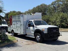 A white 2013 Ford F-550 truck with a flatbed and a silver utility body featuring multiple compartments and a red hydraulic lift mounted at the rear