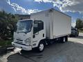 A white 2016 Isuzu NPR box truck with a large cargo area and orange lights on top parked on a concrete surface