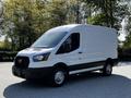 A white 2021 Ford Transit van with a black front grille and large side mirrors, featuring a cargo area and sitting on a flat surface