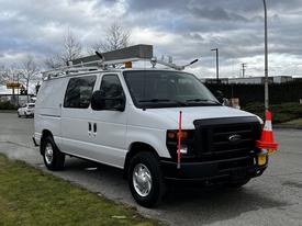 A white 2011 Ford Econoline van with a roof rack and traffic cones placed on the front and rear bumpers