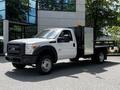 A white 2016 Ford F-550 with a flatbed and enclosed utility box on the back features amber lights on the roof and is parked in front of a modern building