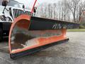 A large orange and black snow plow blade positioned upright in the foreground with the word "PLOW" written on it