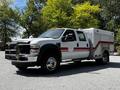 A 2009 Ford F-550 truck with a white body, red stripes, and a utility box on the back is parked in a gravel area with trees in the background