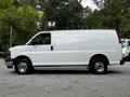 A 2023 GMC Savana cargo van in white with chrome wheels and no side windows parked on a gravel surface viewed from the side