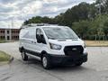 A 2023 Ford Transit van in white with a black lower bumper and roof rack parked on a gray surface