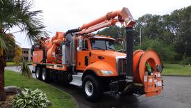 A 2013 Peterbilt 348 vacuum truck in bright orange with a large suction hose and multiple tanks on the side