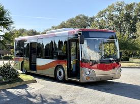 A 2016 Grande West Vicinity bus with a red and silver design parked in a shaded area showcasing its large windshield and modern features