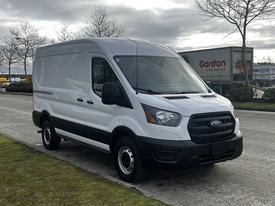 A white 2020 Ford Transit van with a high roof and cargo space visible in the back standing on a street with grass alongside it