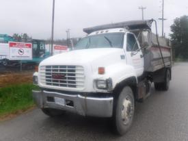 A white 2002 GMC C8500 dump truck with a flatbed and visible wear on the front bumper parked on a roadside