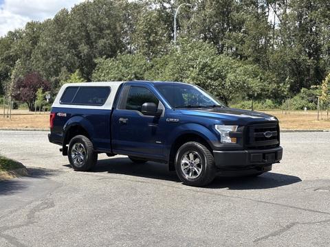 A blue 2015 Ford F-150 with a silver camper shell and chrome wheels parked on a street