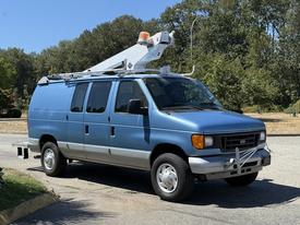 A blue 2006 Ford Econoline van with a mounted equipment rack and safety lights on top parked on a street