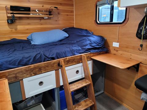 Interior of a 2022 Royal Camper featuring a bed with blue bedding wooden shelves a desk and a small ladder leading to the bed