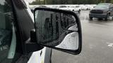 A side mirror of a 2012 Ford Transit covered in raindrops reflecting part of the vehicle's body and nearby vehicles in a parking lot