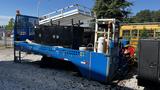 A blue 2000 flat deck truck with an 18-foot platform featuring storage compartments and equipment like propane tanks and tools on the deck