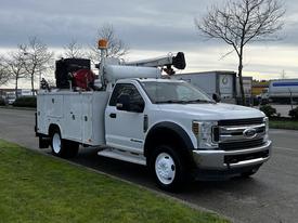 A white 2018 Ford F-550 with a utility body and an elevated work platform mounted on top parked on a street