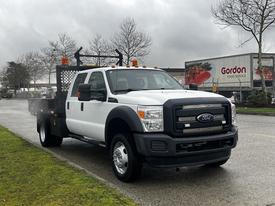 A 2015 Ford F-550 truck with a white body and black front, featuring a flatbed and safety lights on the roof