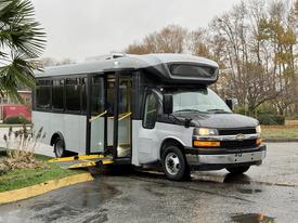 A gray 2019 Chevrolet Express bus with a wheelchair ramp deployed parked at an angle with doors open
