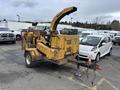 A yellow 2015 Vermeer Corporation BC1200XL wood chipper with a large feeding chute and wheels standing on a parking lot with a small vehicle nearby