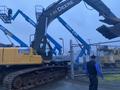 A 2014 John Deere 350G excavator with a long arm and bucket attached, featuring a yellow body and dark tracks, positioned near construction equipment and machinery