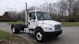 A white 2003 Freightliner M2 106 truck with a single exhaust stack and five-spoke wheels parked with no cargo in the bed