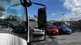 A 2016 Isuzu NPR truck is shown from the driver's perspective with a view through the side mirror and parked cars in the background