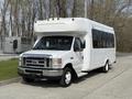 A white 2010 Ford Econoline bus with a high roof and large windows parked on a pavement with trees in the background